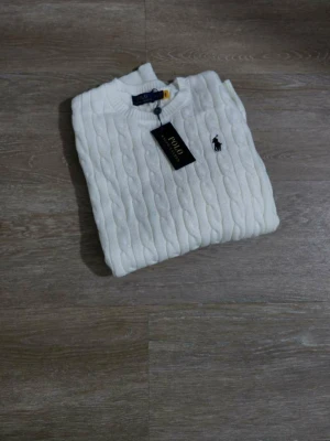 Vit kabelstickad tröja Polo Ralph Lauren - Kabelstickad vit tröja från Polo Ralph Lauren med klassisk rund hals och svart broderad logga på bröstet. Tröjan har ribbade muddar vid ärmslut och nederkant, och är tillverkad i mjukt material som passar perfekt till höst och vinter.