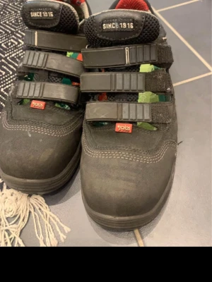 Arbetsskor - Säljer ett par svarta sneakers från Jalas med tre breda kardborreband och vita kontrastsömmar. Skorna har detaljer i grönt och rött på insidan och meshpartier för extra ventilation. Perfekta för dig som vill ha både stil och funktion.