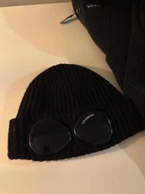 Svart mössa med goggles från C.P. Company - Säljer en svart ribbstickad mössa från C.P. Company med de ikoniska inbyggda goggles framtill. Mössan är i ett mjukt och stretchigt material som sitter skönt på huvudet och har en snygg urban vibe. Äkta, köpt på NK