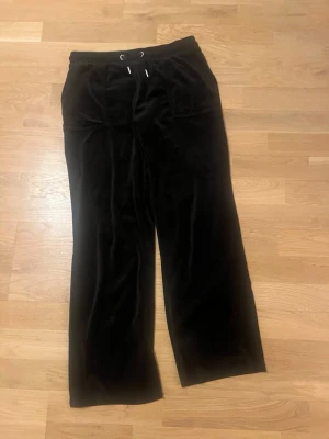 Svarta velourbyxor Juicy Couture - Svarta mjukisbyxor från Juicy Couture i mjuk velour med breda ben och snörning i midjan. Klassisk relaxed passform och fickor både fram och bak. Perfekta för en chill och trendig look.Använd en gång.