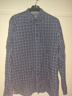 Blårutig skjorta Tommy Hilfiger XL - Snygg blårutig skjorta från Tommy Hilfiger i modellen New York Fit. Skjortan har klassisk krage, långa ärmar och knäppning framtill. Tillverkad i bomull och passar perfekt för dig som gillar stilren och tidlös design.