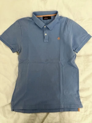 Blå pikétröja från Morris stl M - Snygg blå pikétröja från Morris Stockholm i storlek M. Tröjan har klassisk krage, två vita knappar och en orange broderad logga på bröstet. Materialet är mjuk bomull som känns skönt mot huden. Perfekt för dig som gillar stilrena och enkla plagg.