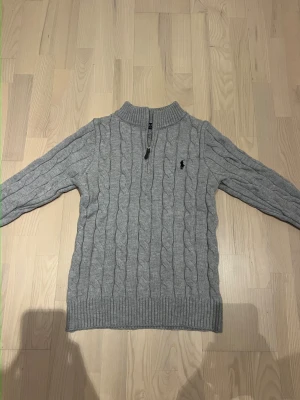 Grå kabelstickad tröja Polo Ralph Lauren - Säljer en grå kabelstickad tröja från Polo Ralph Lauren med half zip och hög krage. Tröjan har lång ärm, ribbade muddar och den klassiska broderade loggan på bröstet. Perfekt för kyliga dagar och enkel att styla till jeans eller kjol.
