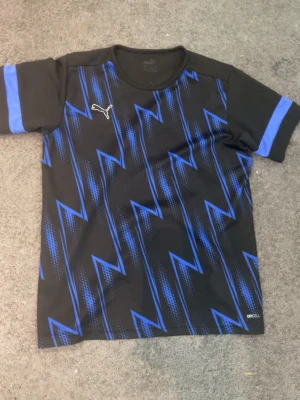 Puma t shirt - Puma t shirt i svart med blixt liknade blå mönster runt om framsidan med en hel svart baksida storlek är 13-14 år för killar en perfekt tränings t shirt