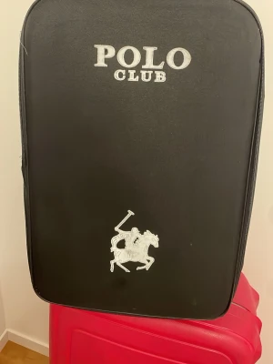 Svart resväska från Polo Club - Säljer en svart resväska från Polo Club med klassisk logga och hästmotiv i vitt. Väskan har en stilren och enkel design med dragkedja runtom. Perfekt för dig som vill resa med stil och hålla koll på dina grejer.