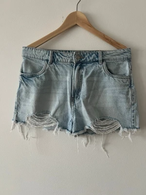 Ljusblå denimshorts från Pimkie - Säljer ett par ljusblå denimshorts från Pimkie med hög midja och råa, slitna kanter samt slitningar framtill. Klassisk femficksmodell med knapp och dragkedja. Perfekta till sommaren för en avslappnad look.