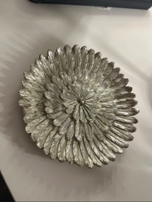 Dekorativ skål i silverfärgad blomdesign - Vacker dekorationsskål med detaljerad blomform och silverfärgad yta. Skålen har ett rikt mönster av lager på lager-blad som ger ett exklusivt och dekorativt intryck. Passar utmärkt som inredningsdetalj, för smycken, nycklar eller andra småföremål. Observera att en liten bit har gått sönder på kanten, vilket syns vid närmare granskning men påverkar inte helhetsintrycket nämnvärt.