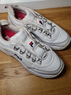 Vita Fila Strada sneakers med metallöglor - Säljer ett par vita Fila Strada sneakers med chunky sula och coola metallöglor för snörningen. Skorna har röda och marinblå detaljer samt Fila-logga på sidan och plösen. Ovandelen är i syntet och textil, insidan är fodrad i rött tyg.