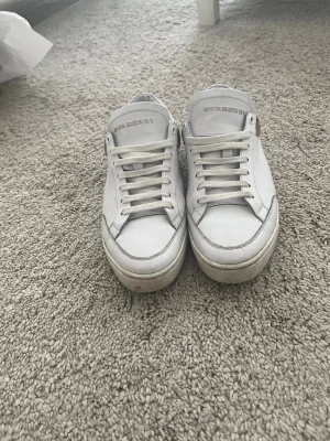 Burberry skor - Säljer ett par vita sneakers från Burberry i skinn med klassiskt rutigt mönster i hälen. Skorna har snörning och rund tå samt en platt sula. Perfekta för dig som vill ha en clean look med en lyxig touch. Skorna är äkta då dom är köpta från trendsellers och har kvitto på telefonen från dom. Säljer dom för ett schysst pris, endast skorna följer med. Vid frågor är det bara att höra av sig!