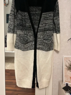 Lång kofta från Crocker i svart och vitt - Vinterns mysigaste kofta! Lång, stickad kofta från Crocker i storlek XS. Koftan har blockfärger i svart, gråmelerat och vitt, två fickor framtill och stängs med svarta knappar. Perfekt för lager på lager under höst och vinter.