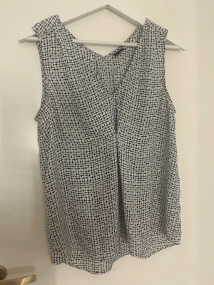 Vit och svart mönstrad blus Gina Tricot - Säljer en vit blus från Gina Tricot i storlek 38 med svart grafiskt mönster. Blusen är ärmlös och har en v-ringad hals. Materialet är lätt och luftigt, perfekt för varmare dagar. Snygg och enkel att matcha med jeans eller kjol.
