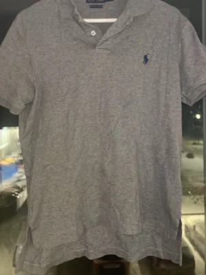 Grå pikétröja från Polo Ralph Lauren - Säljer en klassisk grå pikétröja från Polo Ralph Lauren med blå broderad logga på bröstet. Tröjan har korta ärmar, krage och knäppning med två knappar. Tillverkad i mjuk bomull för en skön känsla.