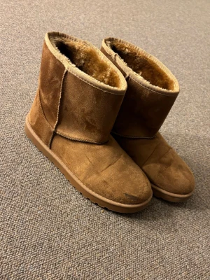 Uggs fejk  - Säljer ett par bruna boots med mjukt pälsfoder på insidan och platt sula. Utsidan är i mocka och de har en rund tå. Perfekta för kalla dagar och har en enkel, klassisk design utan snörning eller dragkedja.