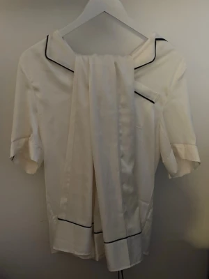 Vit satin pjymas ifrån Primark med svarta detaljer - Säljer en vit pjymas från Primark i storlek 2XS. Pjymas tröjan har korta ärmar, klassisk krage och svarta kantdetaljer längs krage, ficka och ärmslut. Materialet är mjukt och glansigt, troligen polyester. Perfekt för en clean och stilren look. Byxorna är långa och ser exakt ut som toppen.