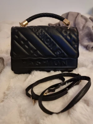 Svart handväska med axelrem - Snygg svart handväska med quiltad yta och texten 'FASHION' i relief. Väskan har ett flätat handtag med gulddetaljer och en avtagbar axelrem. Materialet är skinnimitation och designen har diagonala linjer och stjärnor. Perfekt för dig som gillar trendiga accessoarer.