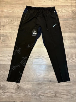 Nike Running Division  - Svarta Nike Running Division träningsbyxor med dragsko i midjan, dragkedjefickor och grafiskt mönster på benen. Byxorna har ventilerande meshfoder och detaljer i mörkblått upptill. Perfekta för löpning eller gymmet.