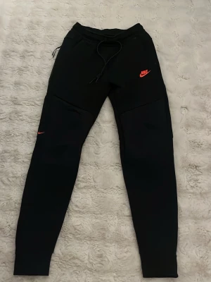 Svarta mjukisbyxor från Nike - Säljer ett par svarta mjukisbyxor från Nike med orange logga på benet och vid fickan. Byxorna har dragsko i midjan och smal passform nedtill. Perfekta för chill eller träning, tillverkade i mjukt material för extra komfort.