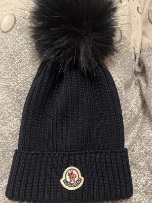 Svart Moncler mössa med pälsboll - Säljer en mörkblå ribbstickad mössa från Moncler med stor fluffig pälsboll på toppen och klassisk Moncler-logga framtill. Mössan är i en enkel, stilren modell och passar perfekt för kalla dagar.