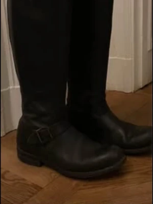 Primeboots  - Säljer ett par svarta höga ridstövlar i skinn med spänne och dekorativ rem upptill och vid ankeln. Stövlarna har dragkedja baktill för enkel på- och avtagning samt en klassisk rund tå. Perfekta för dig som vill ha en stilren och tidlös look.