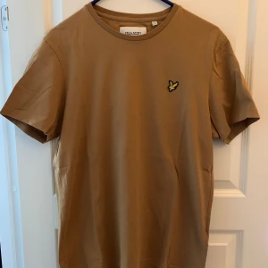 Beige t-shirt från Lyle & Scott - Snygg beige t-shirt från Lyle & Scott med klassisk rund hals och ikonisk gul logga på bröstet. Tillverkad i mjuk bomull och har en enkel, clean look som passar till det mesta. Använd 1 gång så nyskick! 