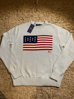 Ljusblå stickad tröja Polo Ralph Lauren - Ljusblå stickad tröja från Polo Ralph Lauren med amerikansk flagga i rött, vitt och blått på bröstet samt RL-broderi. Tröjan har rund halsringning, ribbade muddar och är långärmad. Perfekt för dig som vill sticka ut med en klassisk preppy stil.