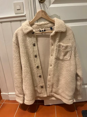 Beige teddyjacka från GANT - Säljer en beige teddyjacka från GANT med mjukt, fluffigt material och klassisk krage. Jackan har knappar framtill och en bröstficka. Perfekt för dig som vill ha en mysig och trendig look under kyliga dagar. I helt nytt skick!