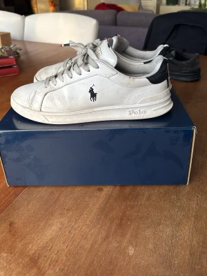 Vita sneakers från Polo Ralph Lauren - Säljer ett par klassiska vita sneakers från Polo Ralph Lauren med marinblå och röda detaljer på hälen. Skorna har snörning, rund tå och Polo-logga på sidan samt guldtext bak. Tillverkade i skinn med textildetaljer. Perfekta till jeans eller shorts.