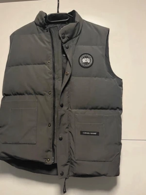 Mörkgrå dunväst från Canada Goose - Säljer en mörkgrå dunväst från Canada Goose med hög krage och två stora fickor framtill. Västen har diskret logga på bröstet och stängs med både dragkedja och tryckknappar. Perfekt för lager-på-lager och riktigt snygg till höst- och vinterlooken.