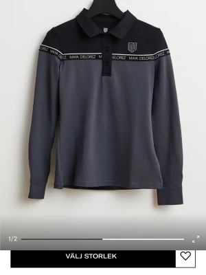 Grå långärmad tröja Maya Delorez - Snygg långärmad tröja från Maya Delorez i mörkgrått med svart axelparti och krage. Tröjan har tryckt logoband över bröstet och knappar vid halsen. Perfekt för dig som gillar sportig och stilren design.