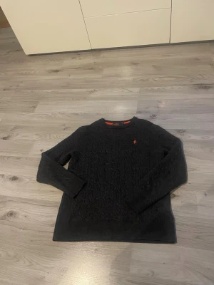 Mörkgrå kabelstickad tröja Morris - Säljer en mörkgrå kabelstickad tröja från Morris i 100% merinoull. Tröjan har rund hals, lång ärm och en liten orange broderad logga på bröstet. Perfekt för kyliga dagar och riktigt skön att ha på sig.