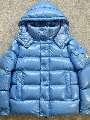 Blå Moncler pufferjacka med huva - Säljer en riktigt fet blå pufferjacka från Moncler. Jackan har glansig yta, stor huva och hög krage. Feta detaljer som logga på ärmen, dragkedja med Moncler-zip och tryckknappar. Perfekt för kalla dagar när du vill se cool ut.