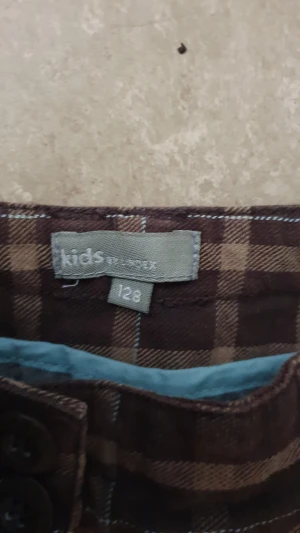 Ankellånga byxor - Snygga bruna ankellånga byxor från Lindex i rutigt mönster. Byxorna har knappar vid benslut och bakfickor med lock och knapp. Perfekt för dig som gillar en chill och unik stil.