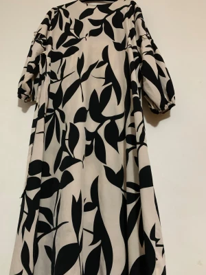 Beige långklänning med svart mönster - Säljer en beige långklänning med svart grafiskt bladmönster. Klänningen har voluminösa trekvartsärmar med resår vid ärmslut och en rak, luftig passform. Materialet känns lätt och flowy, perfekt för dig som gillar statement-plagg med coola prints.