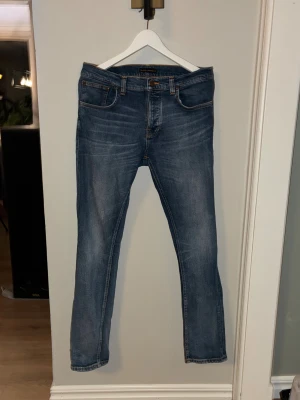  Nudie Jeans W33 L32 - Säljer ett par klassiska blå jeans från Nudie Jeans med orangea sömmar och snyggt broderad bakficka. Modellen har rak passform och normal midja, tillverkad i slitstarkt bomullsmaterial. Ikonisk Nudie-logga på läderpatch baktill.