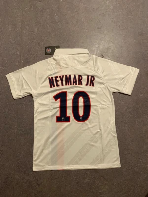 PSG Neymar Jr 10 fotbollströja Nike - Snygg PSG fotbollströja med Neymar Jr och nummer 10 på ryggen. Tröjan är vit med en röd och marinblå rand på framsidan, broderat Nike-märke och PSG-logga. Klassisk krage och korta ärmar. Tillverkad i lätt och ventilerande polyester, perfekt för match eller träning.