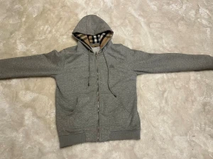 Grå Burberry hoodie med rutig foder - Säljer en grå hoodie från Burberry med klassiskt rutigt foder i huvan och insidan. Tröjan har dragkedja, snörning vid huvan och två fickor fram. Perfekt för dig som vill ha en stilren och lyxig look med ikonisk Burberry-touch.