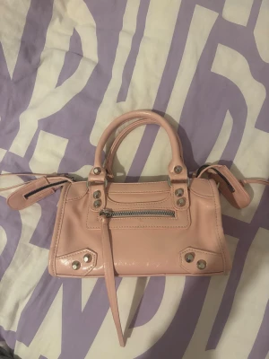 Ljusrosa handväska med silverdetaljer - Snygg ljusrosa handväska i blankt syntetmaterial med silverfärgade nitar och dragkedjor. Väskan har två handtag, en yttre ficka med dragkedja och dekorativa remmar. Insidan är svart med en innerficka med dragkedja. Perfekt storlek för det viktigaste.