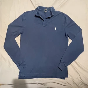 Marinblå långärmad pikétröja Polo Ralph Lauren - Snygg marinblå långärmad pikétröja från Polo Ralph Lauren med klassisk krage och två knappar. Tröjan har det ikoniska broderade polospelar-loggan i vitt på bröstet och är gjord i mjuk bomull. Storlek M. Skick 10/10 – Som Ny. Nypris ca 1200-1600 kr. ✅ Äkta vara | 📩 Öppen för frågor | 🚚 Skickas inom 24 timmar 