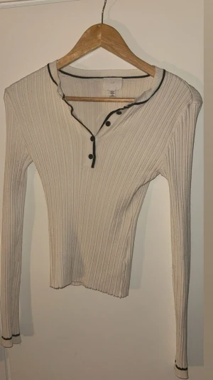 Beige ribbad långärmad topp H&M - Säljer en beige ribbad långärmad topp från H&M med svarta detaljer längs krage och ärmslut. Toppen har v-ringad hals med svarta knappar och en slim passform som sitter snyggt. Perfekt för dig som gillar stilrena och enkla plagg. Ej synligt använd, använd nån gång..