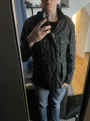 Field jacket - Svart parkajacka med fyra stora fickor framtill och snygga guldfärgade knappar. Jackan har dragkedja och hög krage, perfekt för dig som gillar en clean och funktionell stil. Materialet är slitstarkt och känns lätt mot kroppen. Passar dig som vill ha en jacka med lite edge.