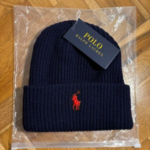 Mörkblå ribbstickad mössa Polo Ralph Lauren - Snygg mörkblå ribbstickad mössa från Polo Ralph Lauren med klassisk röd broderad logga framtill. Tjockt uppvik och stilren design som passar perfekt till kalla dagar. Materialet är mjukt och värmande, perfekt för vinter och höst.