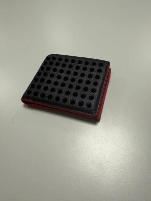 Christian Louboutin plånbok - Snygg plånbok från Christian Louboutin i svart skinn med röda detaljer. Framsidan har coola svarta nitar i ett prickigt mönster och insidan är helröd med plats för kort och mynt. Perfekt för dig som gillar statement-accessoarer. KAN GÅ NER LITE VID SNABB AFFÄR