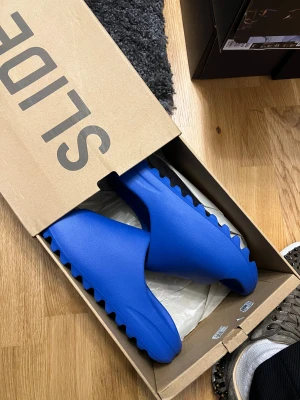 Blå Yeezy Slide sandaler - Säljer ett par blå Yeezy Slide sandaler med chunky, räfflad sula och öppen tå. De har en clean och modern design i syntetmaterial som är både lätt och bekvämt. Perfekt för dig som gillar streetwear och vill sticka ut med en fet färg på fötterna. 43 fit 41-42