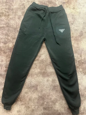 Svarta mjukisbyxor från Prada - Svarta mjukisbyxor från Prada med klassisk logga på vänster lår. Byxorna har snörning i midjan och ribbade muddar vid bensluten. Tillverkade i mjukt bomullsmaterial för maximal komfort och stil. Perfekta för chill eller streetwear.