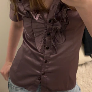 Lila glansig blus med volanger från Shesay - Supersnygg lila blus från Shesay med glansigt tyg och volanger längs v-ringningen. Knappar framtill och korta puffärmar ger en cool och fin vibe. Bara använd ett fåtal gånger och inga skavanker!