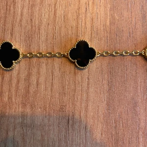 Van Cleef & Arpels Alhambra armband - Exklusivt Alhambra armband från Van Cleef & Arpels med fem svarta fyrklöver i onyx och guldfärgad kedja. Ikonisk design med detaljerad pärlkant runt varje klöver. Perfekt statement piece för dig som gillar lyxiga accessoarer. Skicka prisförslag