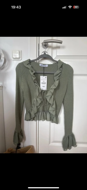 Grön croppad volangtop från Zara - Snygg ljusgrön croppad topp från Zara med volanger längs halsringningen och nederkanten. Ribbad stickad struktur, långa ärmar med volangdetaljer och knyt framtill. 