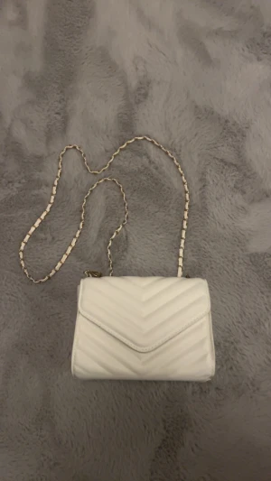Vit quiltad kuvertväska med kedja - Snygg vit kuvertväska med quiltad design och guldfärgad kedja som axelrem. Väskan har ett stilrent mönster i V-form på framsidan och är tillverkad i skinnimitation. Perfekt för att lyfta din outfit med en elegant touch.
