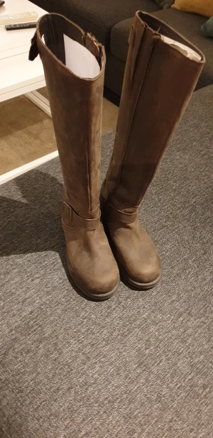 Bruna höga skinnstövlar med spänne - Snygga höga stövlar i brunt skinn med rund tå och platt sula. De har dekorativa spännen vid ankeln och dragkedja på insidan för enkel på- och avtagning. Perfekta för höst och vinter, passar till både jeans och kjol.