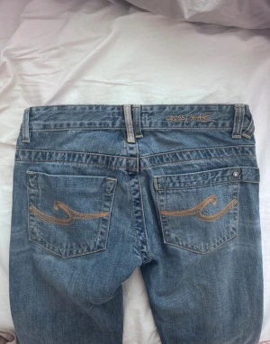 Blå Cross Jeans low waist bootcut - Säljer ett par blå jeans från Cross Jeans med snygg broderad detalj på bakfickorna och röd knapp framtill. Modellen är lågmidjad med bootcut och klassisk femficksdesign. Jeansen har en chill passform och är gjorda i mjukt denimtyg.